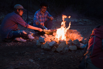 Happy friends camping enjoying bonfire in nature at night, Partying in camping, Asian Young people celebrating during summer vacation, summertime and travel activity. Selection focus to bonfire.
