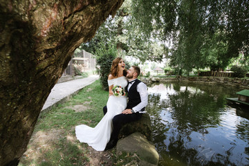 Fototapeta premium Stylish bride and groom sitting near Lake and hugs in wedding day. Holidays, vacation, love, wedding concept. Place for text