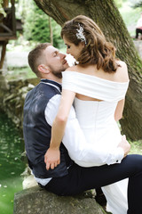 elegant stylish groom with his happy gorgeous brunette bride with curves hair and wedding bouquet are kissing on the background of a lake. Holidays, vacation, love, wedding concept. Place for text