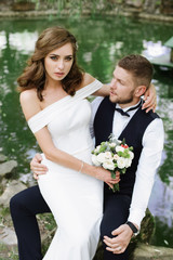elegant gentle stylish groom with his happy gorgeous brunette bride with curves hair and wedding bouquet . Holidays, vacation, love, wedding concept. Place for text hugs on the background of a lake