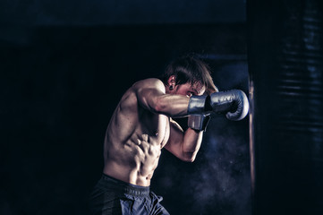Fototapeta na wymiar Strong and muscular guy practicing punching the boxing bag. Moody environment. Edgy edit style.
