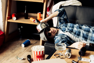 handsome man holding teapot near head while lying on sofa in messy living room
