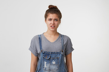 Young beautiful attractive girl dressed in a denim overall looks upset displeased unpleasantly surprised, angry, saw something disgusting.
