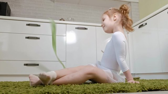 A happy little girl in a white gymnastic swimsuit trains, dances with a ribbon for rhythmic gymnastics, jumps and performing professional exercises.