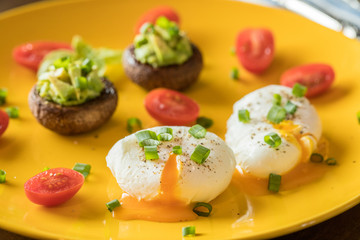 Poached eggs with avocado stuffed mushrooms, tomatoes, and green onions on a cheery yellow plate