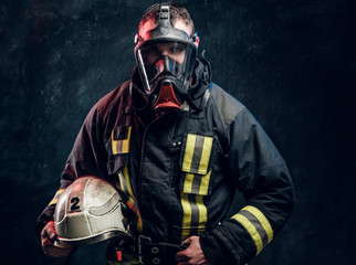 Brutal fireman in gas mask holding his helmet  and looks into camera on black background in studio