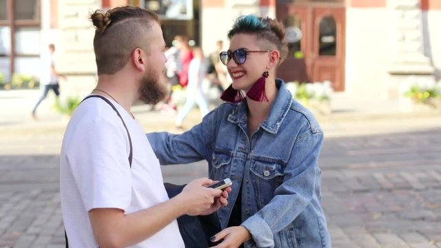 hipster guy with a beard with a smartphone in his hand meets a girl. hipster kisses when meeting and shows something on the smartphone.