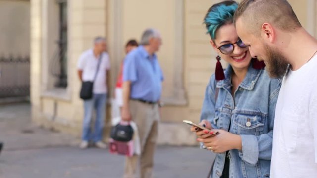Beautiful hipster Caucasian girl in jeans jacket with a smart phone in her hands shows something to a hipster guy in her smartphone both laugh the camera moves back