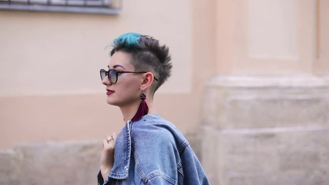 beautiful caucasian girl with hipster hairstyle in a dress in sun-protection glasses holding a jeans jacket goes and the camera moves from right to left