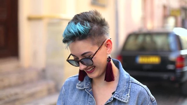 beautiful caucasian girl with hipster hairstyle in sun-glasses in jeans jacket in hands smartphone and coffee laughing goes and looks into smartphone