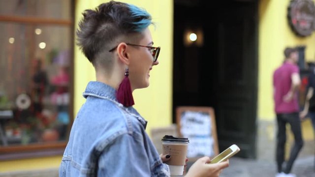 hipster girl with a smartphone in her hands and with coffee, goes back to the camera, passing people looks into the smartphone