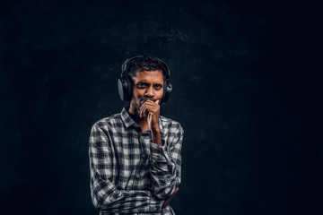 Portrait of a handsome Indian man wearing a checkered shirt listens to music in wireless headphones with thoughtful look standing in studio against the background of the dark wall.