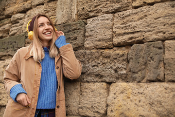 Young woman with headphones listening to music near stone wall. Space for text