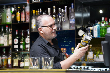 Elderly bartender is holding a bottle of whiskey. The barman examines a bottle of whiskey. The bartender pours whiskey to the client in the hotel bar. The concept of service. 