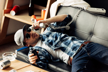 handsome man in sunglasses and cap lying on sofa and talking on retro phone in living room