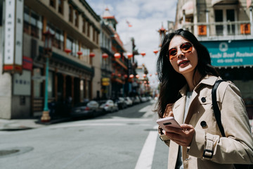 Fototapeta na wymiar Happy young woman in sunglasses enjoying weekends spending time outdoors holding smartphone for chatting. cheerful girl laughing on street looking away mobile phone in hand walking in chinatown.