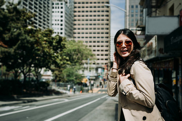 young asian lady backpacker in sunglasses face camera smiling charming. girl tourist laughing carrying bag standing on street with trees in park in spring. happy woman on road in busy shopping area.