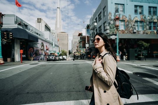 Beautiful asian travel woman wearing fashionable sunglasses holding smart phone in hand. young girl backpacker walking on street in chinatown on sunny day. tall tower Transamerica Pyramid in back.