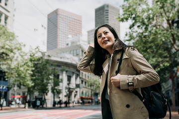 Obraz premium asian traveler woman smiling cheerful standing on street with green trees on sidewalk in spring. young girl local lifestyle go to work in morning san francisco usa. female backpacker sightseeing city
