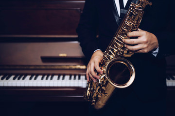 close up of Young Saxophone Player hands holding alto sax musical instrument with piano background in dark room, vintage tone, can be used for music background, copy space 