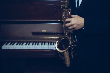 close up of Young Saxophone Player hands holding alto sax musical instrument with piano background in dark room, vintage tone, can be used for music background, copy space 