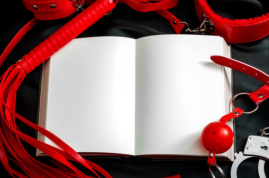 Sexual kink and BDSM concept theme with an open book or notebook with blank white pages sounded by handcuff, whip, flogger, collar and leash, ball gag and other bondage devices with copy space