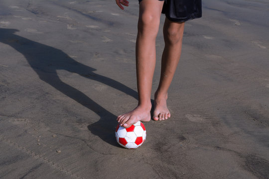 El ni&ntilde;o est&aacute; pisando el bal&oacute;n de f&uacute;tbol sobre la arena de la playa.