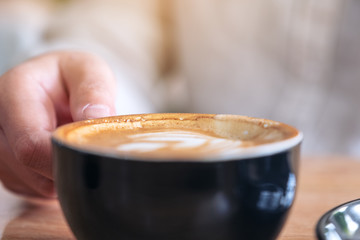 Closeup image of a hand holding a cup of hot coffee with latte art on wooden table