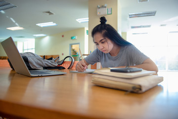 Asian female students learning from education network with laptop and making notes writing down information in the library. Learning and preparing for university exam.