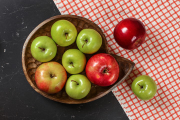 Top down view, green and red shiny apples on wooden carved bowl, red gingham tablecloth and dark marble board under.