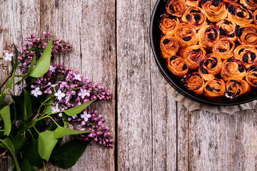 Homemade mini cherry rolls in baking form with bouquet of lilac on vintage wooden table. Bun cake with cherry filling