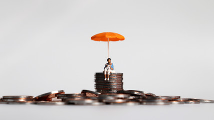 A miniature woman holding a baby sitting with an orange umbrella on a pile of coins.