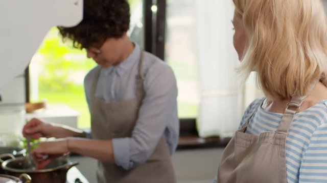 Tilt up of middle-aged blonde woman making buns from dough and talking to young woman with short curly hair mixing soup with spoon and salting it