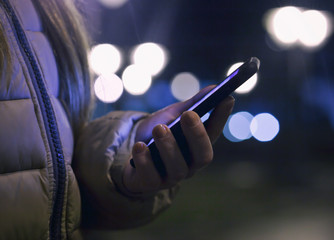 The young woman holds her phone and read some text in the evening city in winter