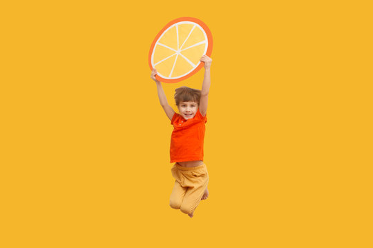 Funny Kid With An Orange Segment From A Cardboard Jump On A Yellow Background. Healthy Food. Concept Summer Lifestyle. 