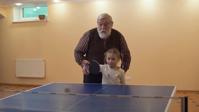 Grandfather teach his granddaughter to play table tennis, standing near her. Little girl beats off the ball. Leisure of happy family