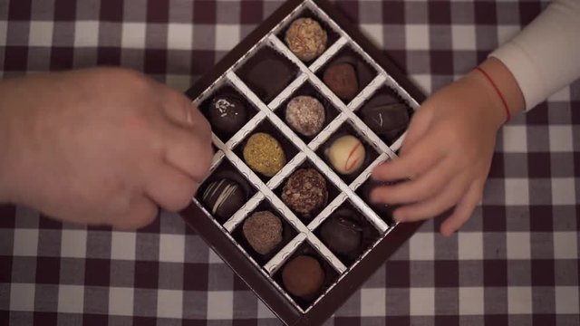 The box of candies stands on the table. Four hands taking sweets one by one. Children and grandparents eat chocolate at home. Close up.