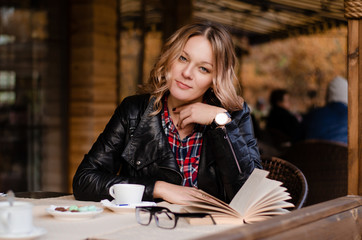 Attractive young woman sitting alone at the table with book in street cafe, drinking coffee or tea.  Relaxing in cafe during free time