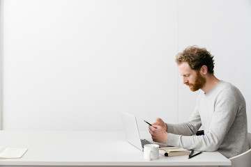 A thirty something man with a red beard and curly hair in a grey pull over is looking on his smart phone while working on his laptop in a clean white empty office setting.