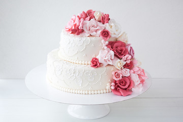Two-tiered white wedding cake decorated with pink flowers on a white wooden background.