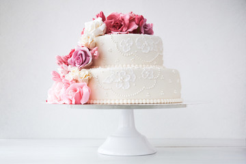 Two-tiered white wedding cake decorated with pink flowers on a white wooden background.