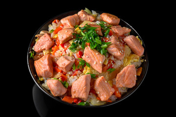 A plate of delicious rice with grilled salmon pieces and vegetables: carrots, sweet peppers, greens, spices and sauce on a black background