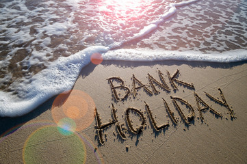 Bank Holiday message handwritten on the smooth sand of an empty beach with an oncoming wave - Powered by Adobe