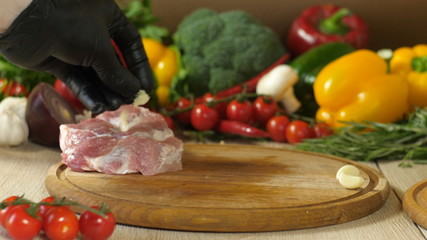 The chef in black gloves marries a piece of raw meat with garlic, which squares with a knife, a branch of rosemary and thyme, the preparation of meat for roasting on the grill, in the background