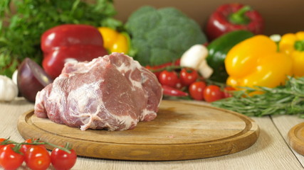 a piece of raw juicy meat on a round wooden board, in the background vegetables greens, broccoli, red pepper, red onion, garlic, cherry tomatoes, sprigs of rosemary and thyme, the background is