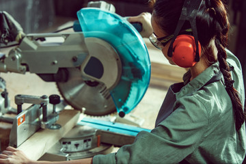 Busy and serious craftswoman grinding timbers with special machine. Beautiful woman wearing safety...