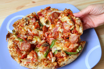 Woman's Hand Picking a Piece of Italian Sausage with Bell Pepper Pizza Served on a Blue Plate