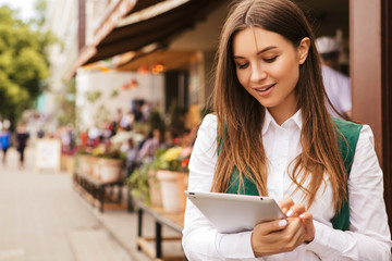 Fototapeta na wymiar Beautiful brunette business woman in white blouse and green jacket working on a tablet in her hands outdoors. Freelancer in european city. Space for text