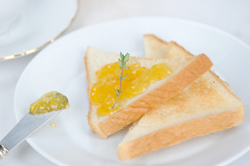 Toasts with orange jam on a white plate. A knife with the remnants of jam lies on the edge of the plate. Toast decorated with a sprig of thyme. Light background. Close-up. Macro shooting.