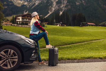 Young woman sitting in the car trunk with suitcases, mountains in the background